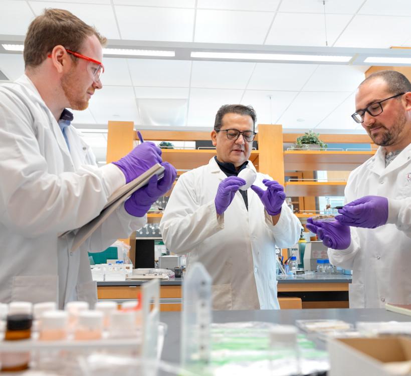 three men in a lab wearing lab coats and purple gloves