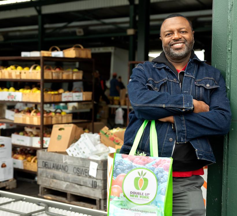 man in a jean jacket smiling at the camera holding a shopping bag with fruit and vegetable stalls behind him