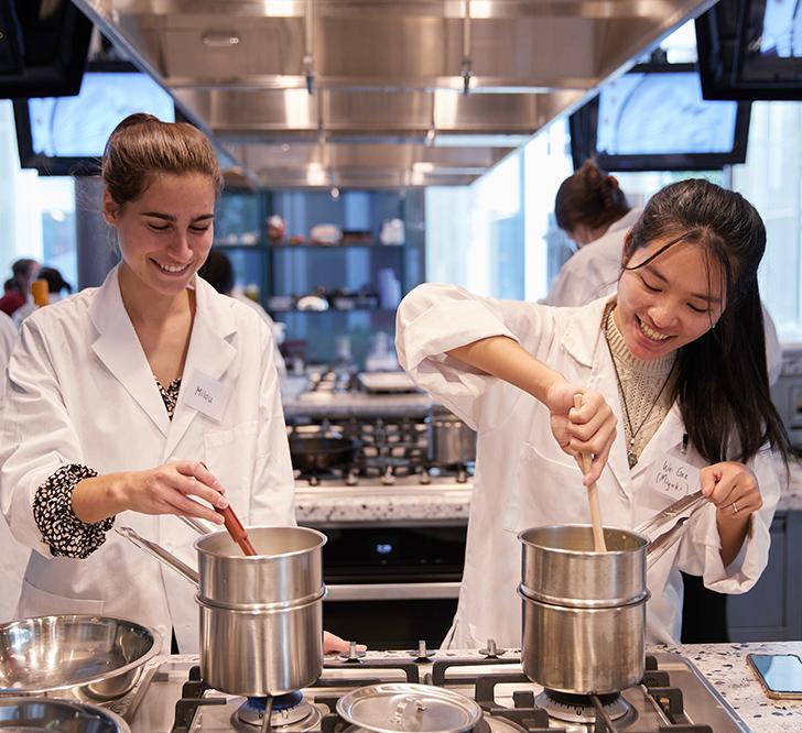 two undergrads in lab coats in an industrial kitchen standing side by side at a stovetop stirring identical pots
