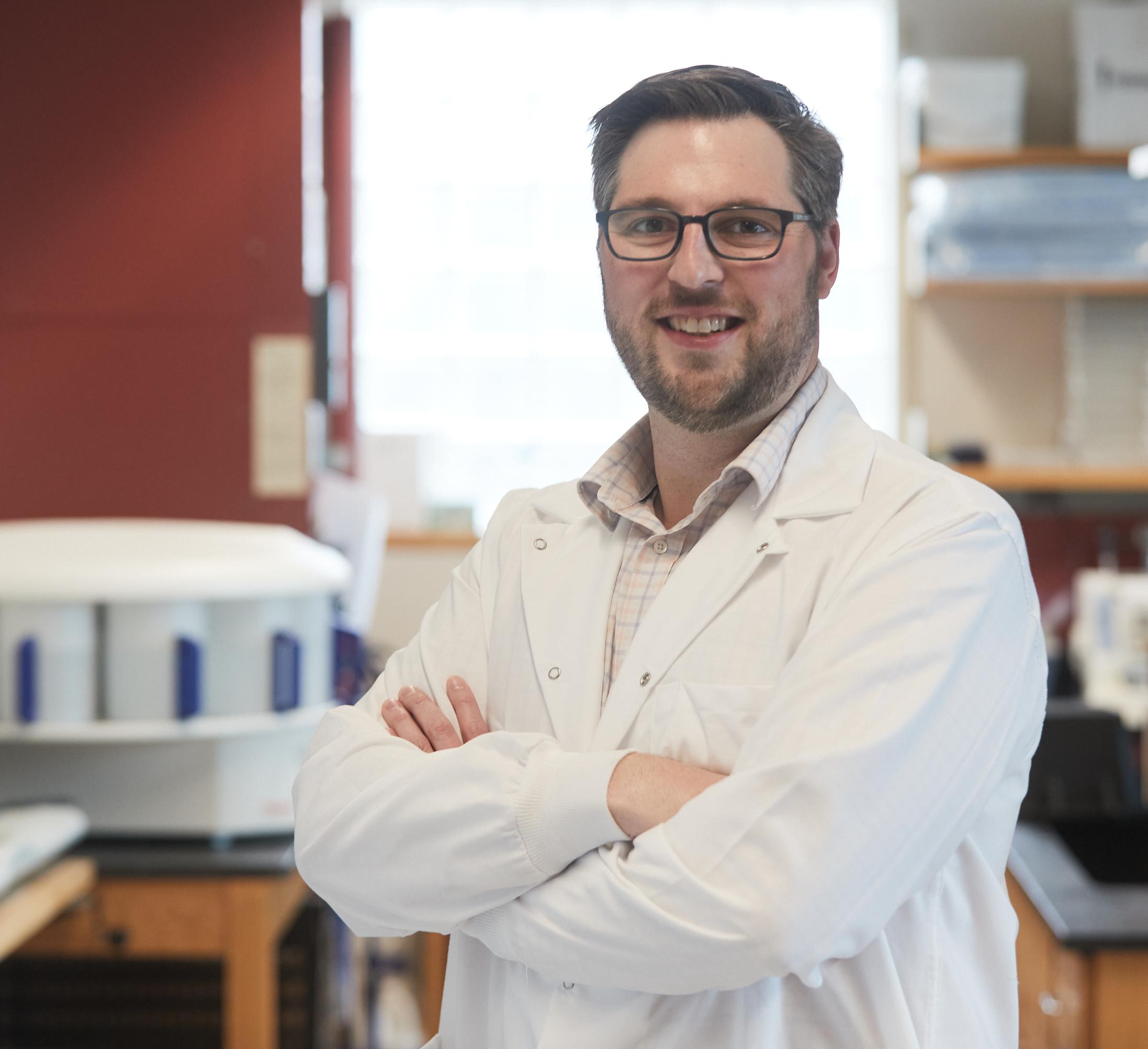 Man in a lab coat standing with crossed arms with a laboratory behind him