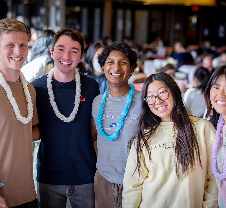 five undergraduate students standing in a dining hall, wearing plastic leis 