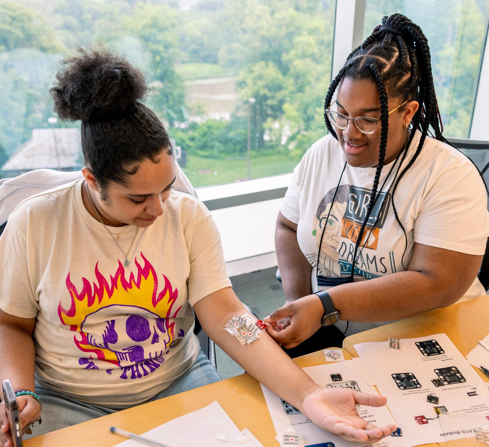 two high school students sitting at a table. one is attaching a sensor to the other's forearm