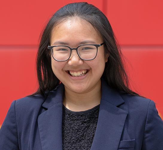 college senior wearing glasses and a blue blazer smiling with a red background