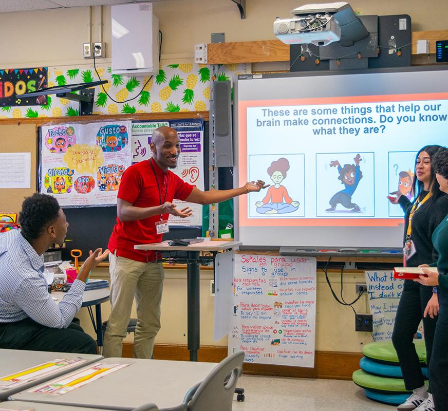 four people stand in an elementary school classroom looking at a presentation slide reading " There are some things that help our brain make connections. Do you know what they are?"