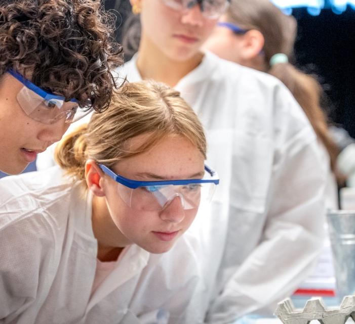 Teens look at an experiment while wearing safety glasses and lab coats