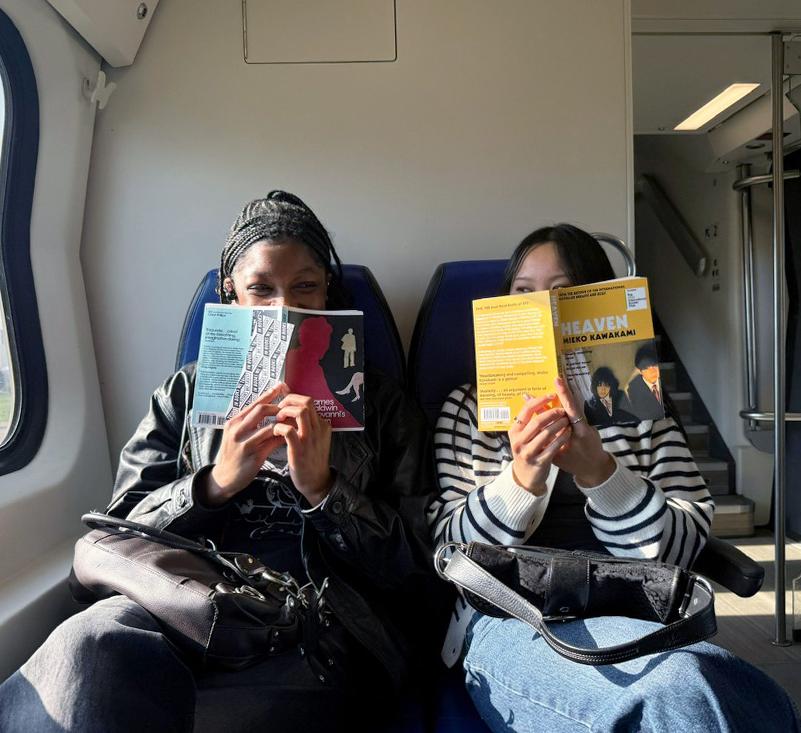 Two women sit on a train in Europe with books in front of their faces 