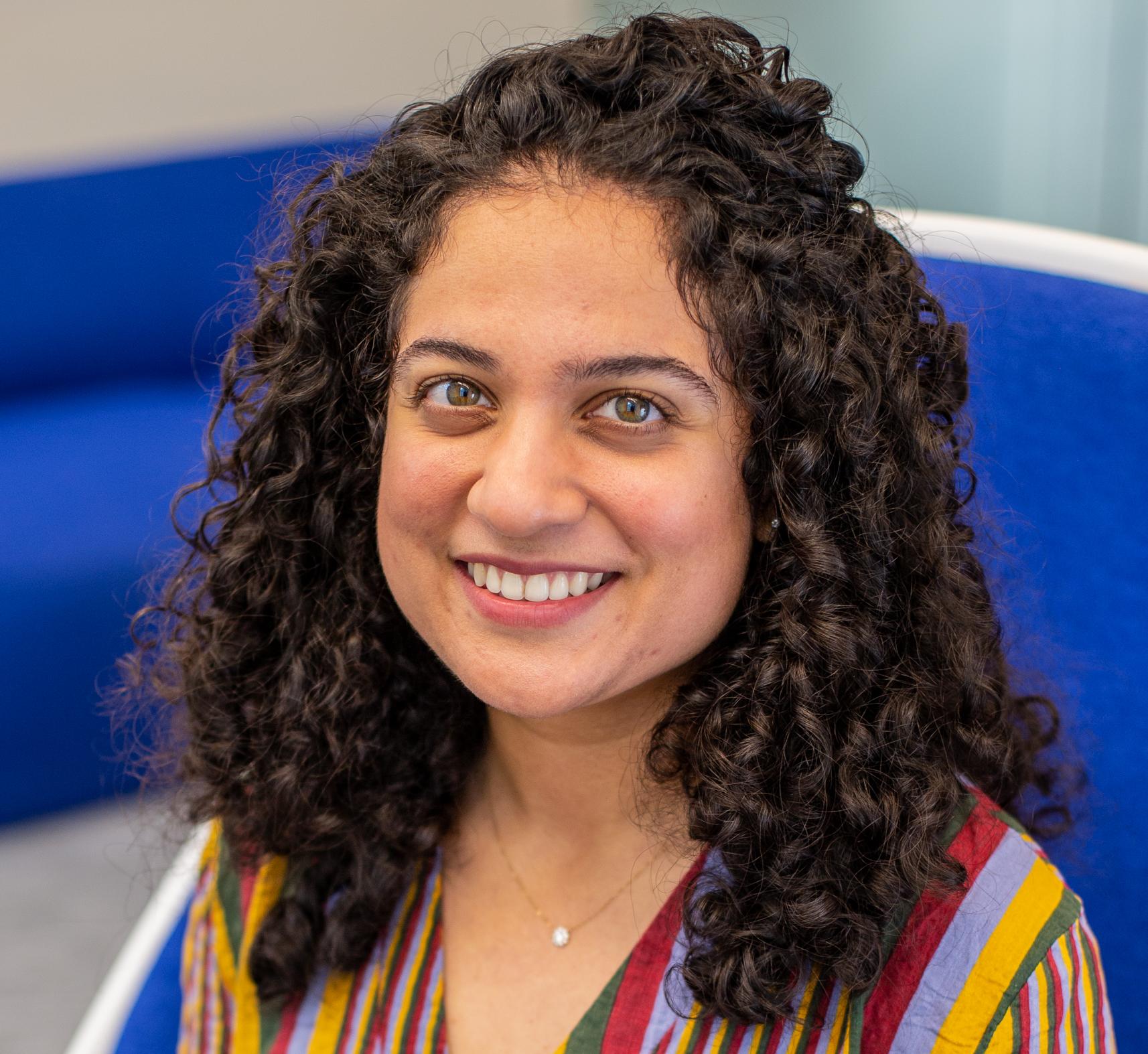 woman with curly dark hair and a brightly patterned shirt sits in a bright blue chair