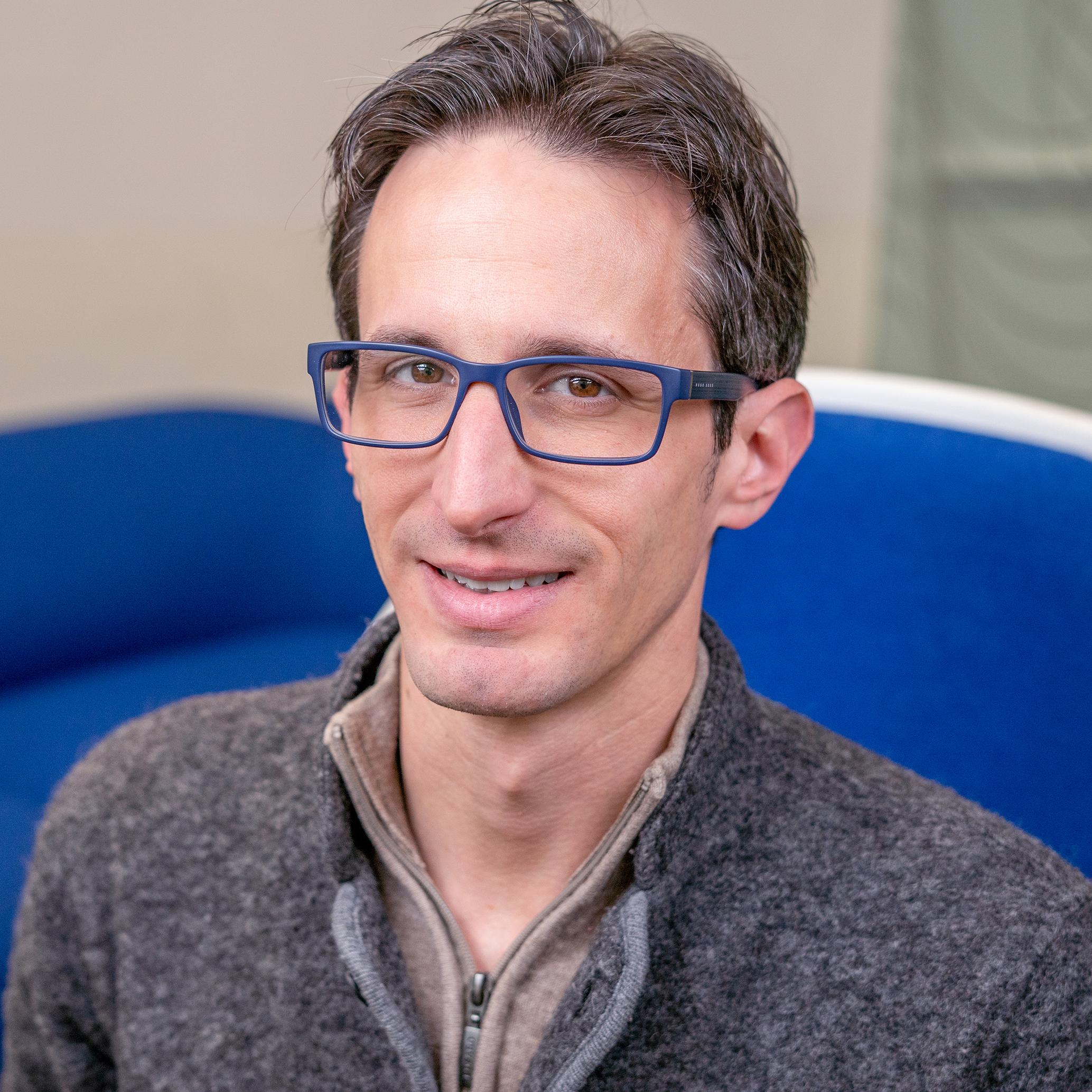 portrait of a man wearing glasses sitting in a bright blue upholstered chair