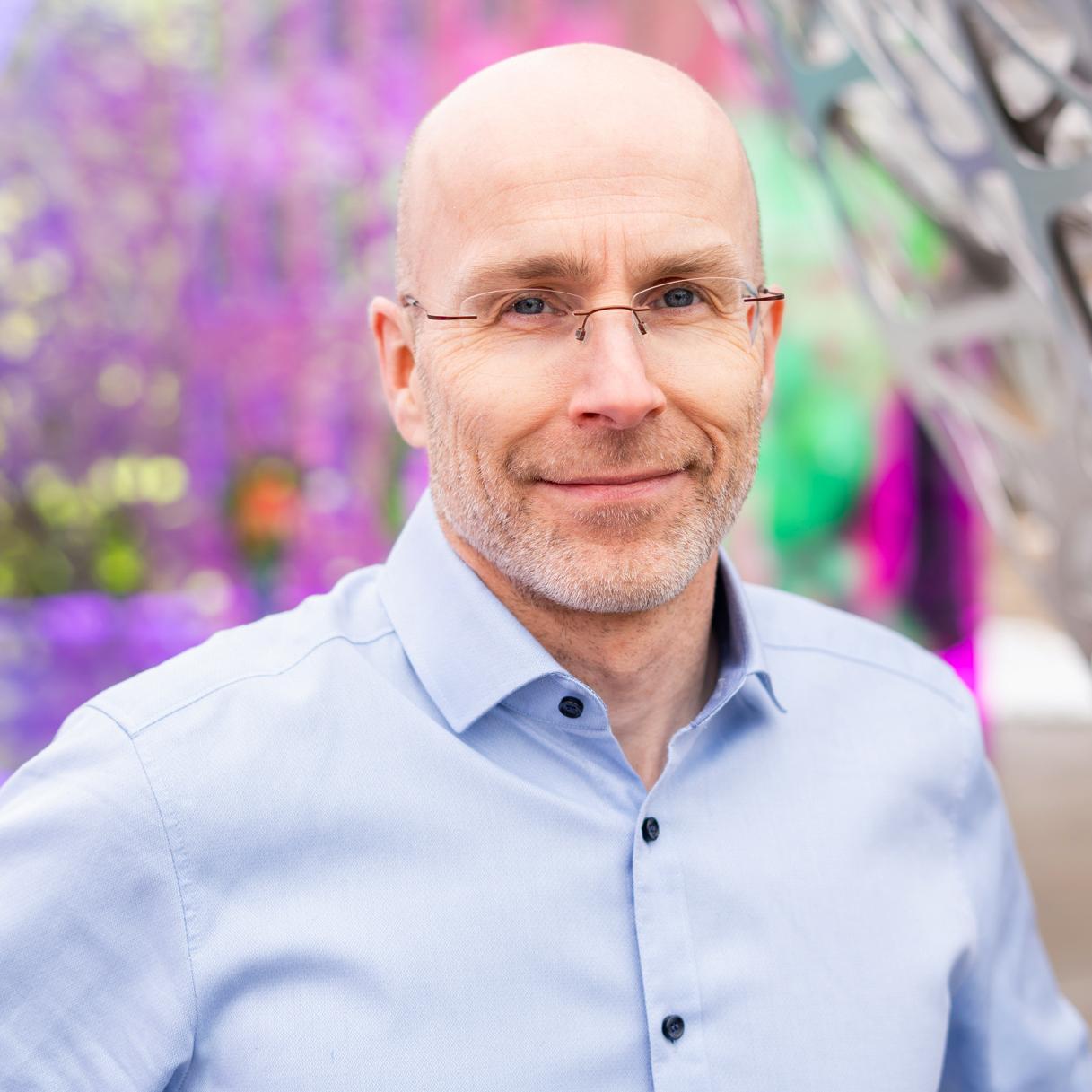 portrait of a man in a blue button down shirt standing in front of a colorful outdoor sculpture