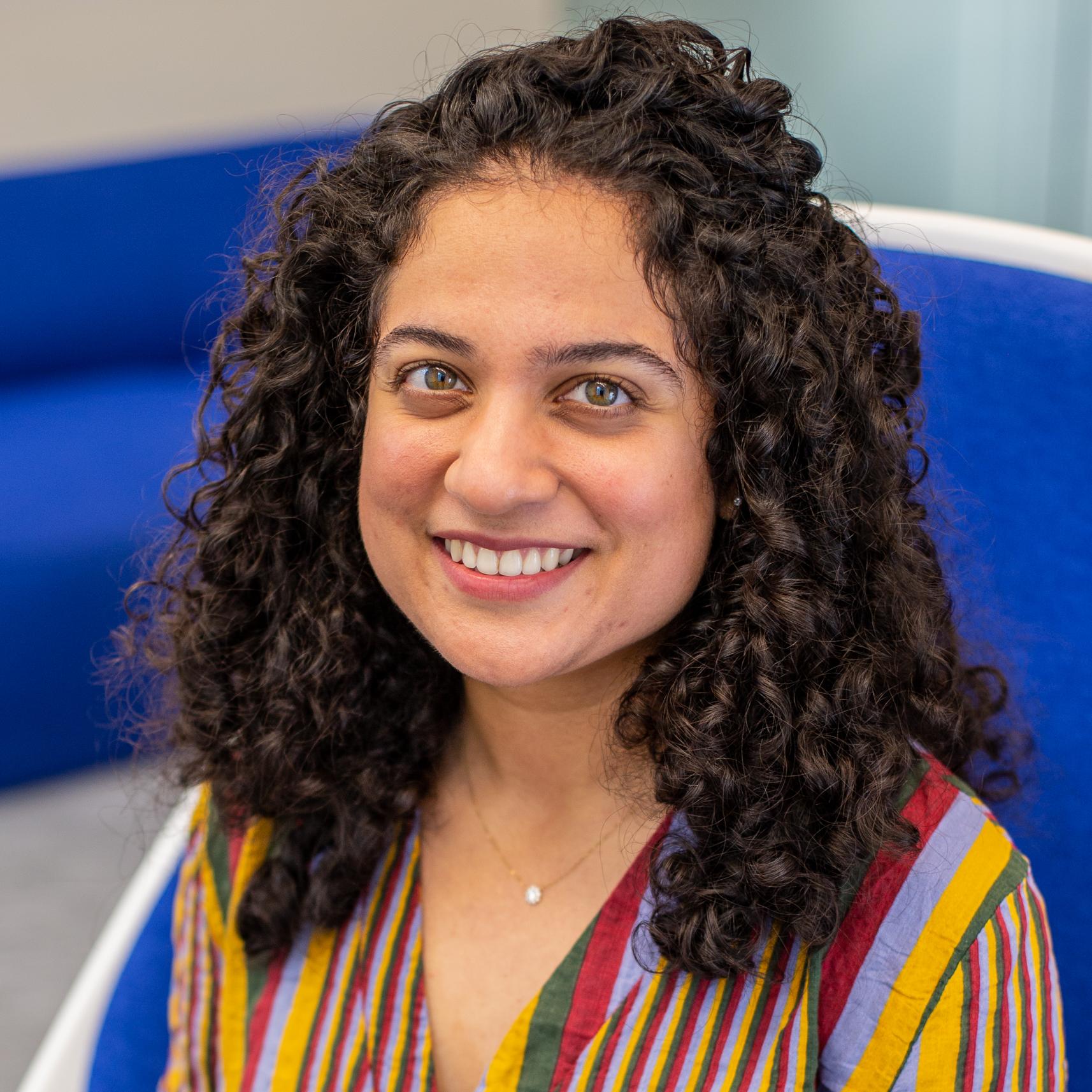 woman with curly dark hair and a brightly patterned shirt sits in a bright blue chair