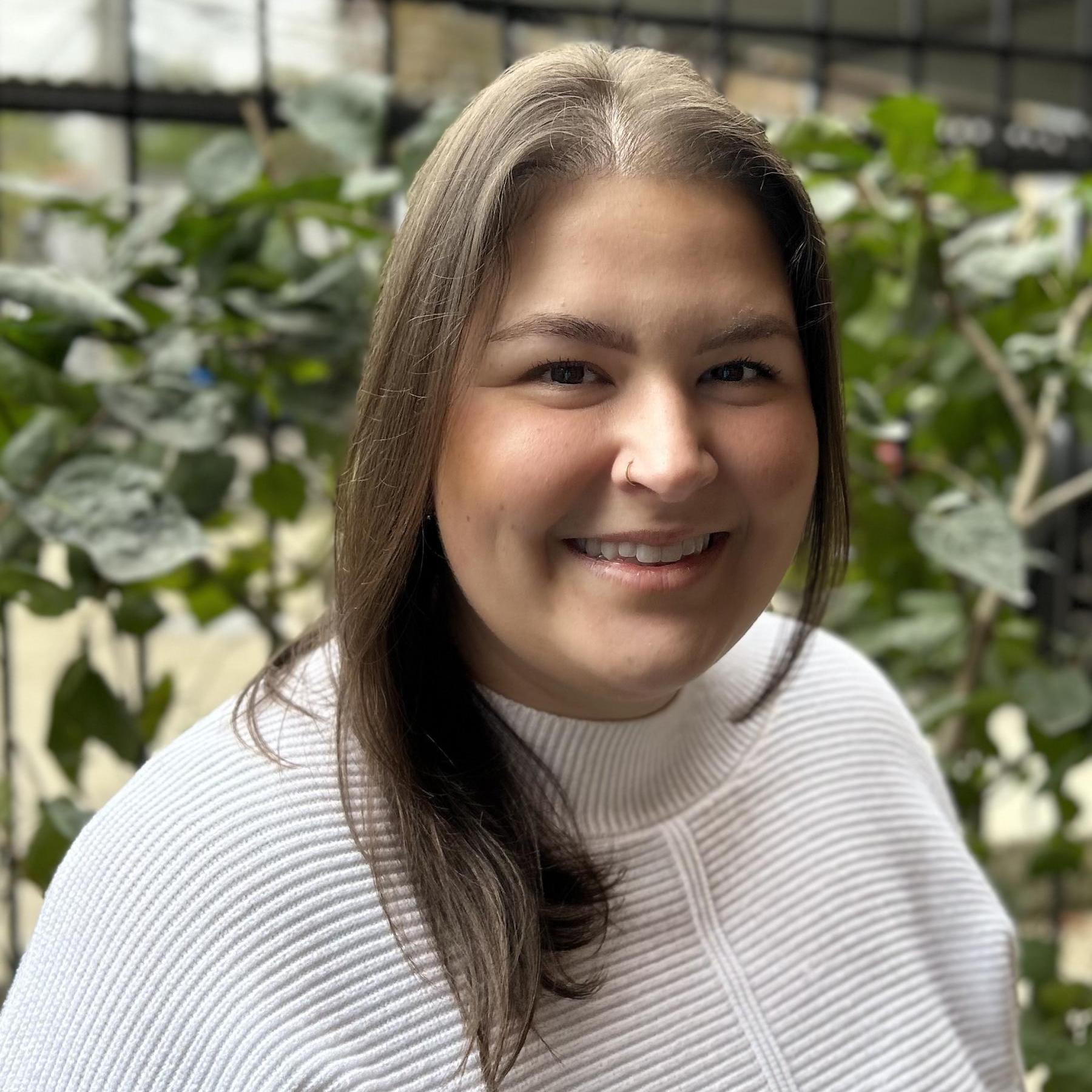 A woman in a white sweater and chest-length brown hair stands in front of plants