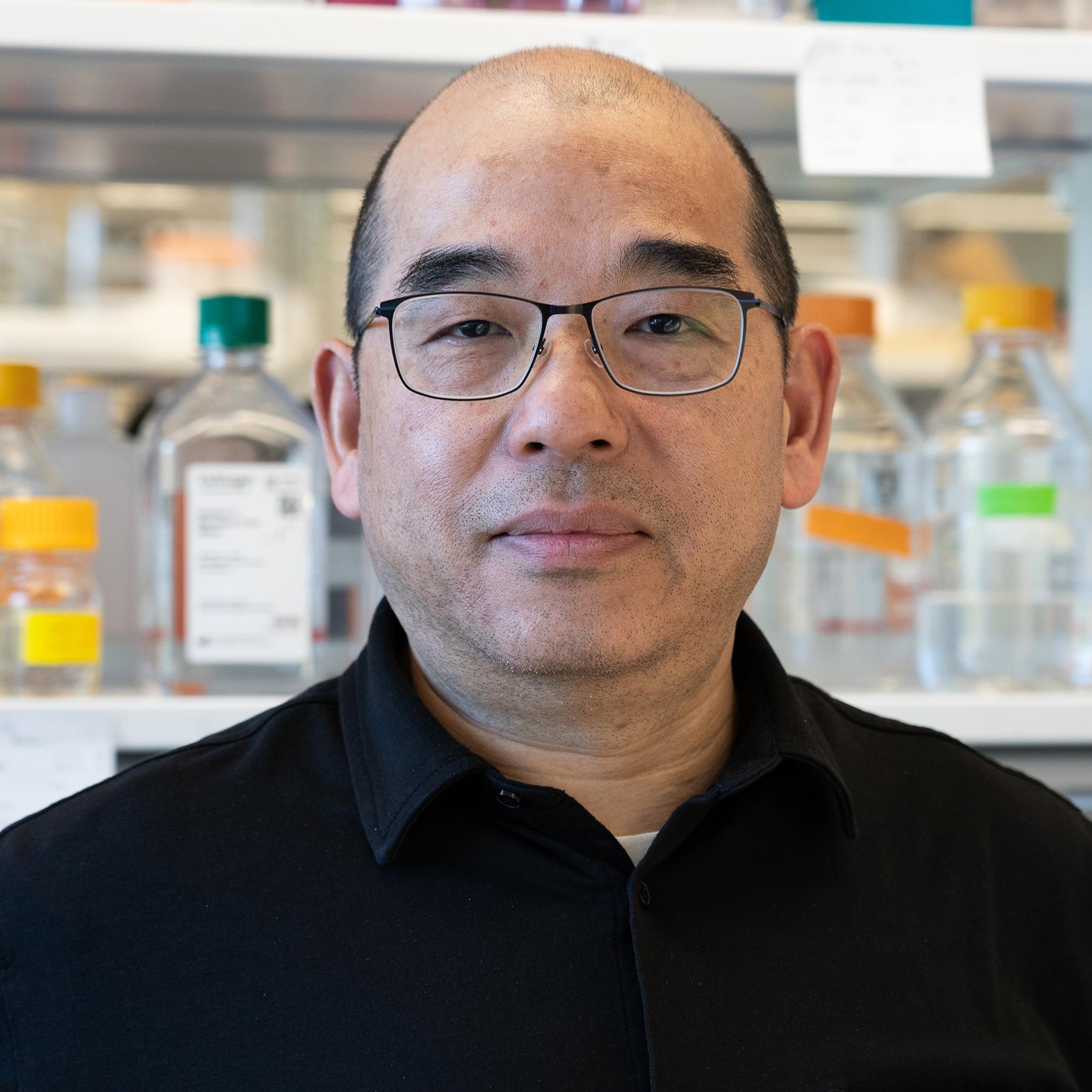 man wearing glasses standing in a lab with shelves of glass bottles behind him