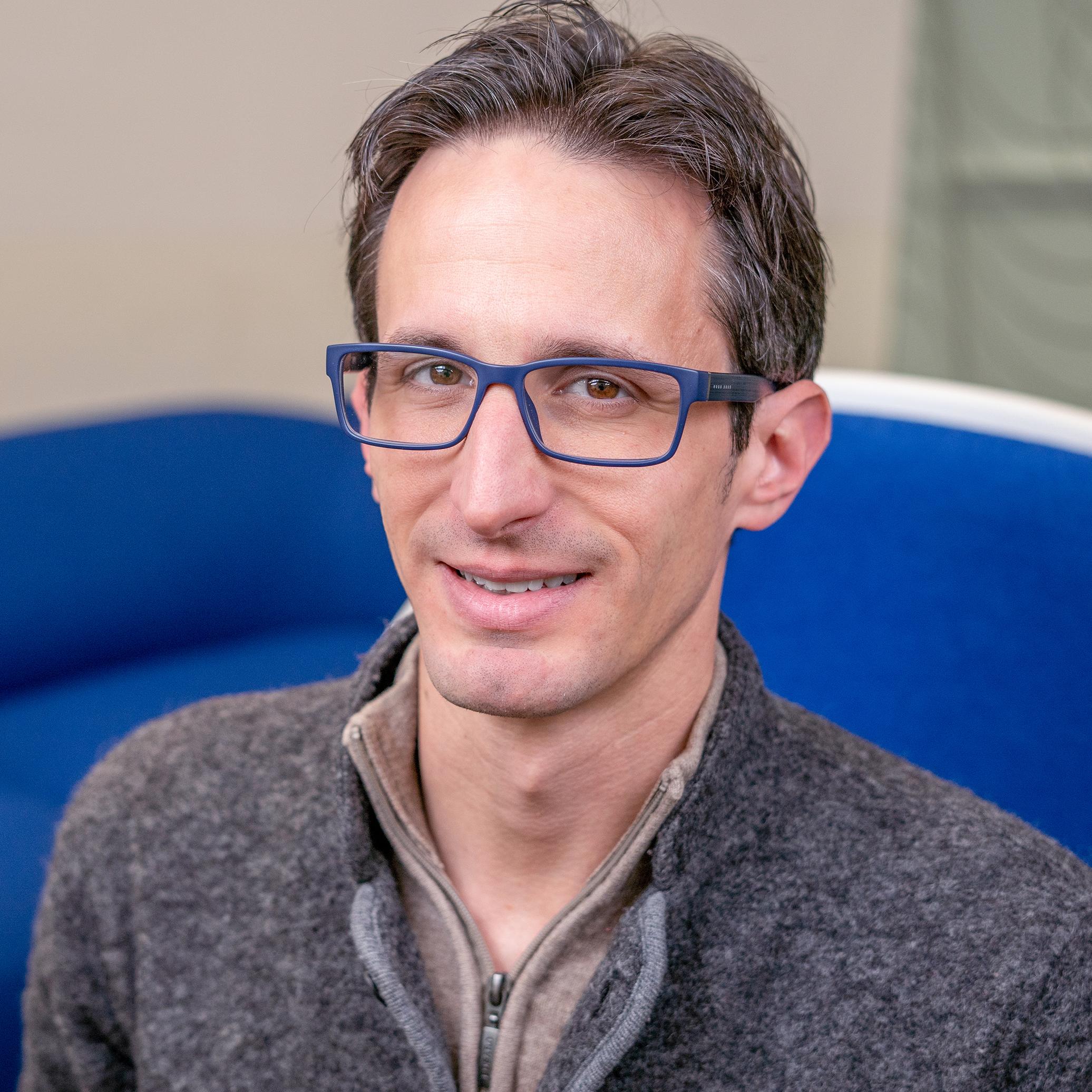 portrait of a man wearing glasses sitting in a bright blue upholstered chair