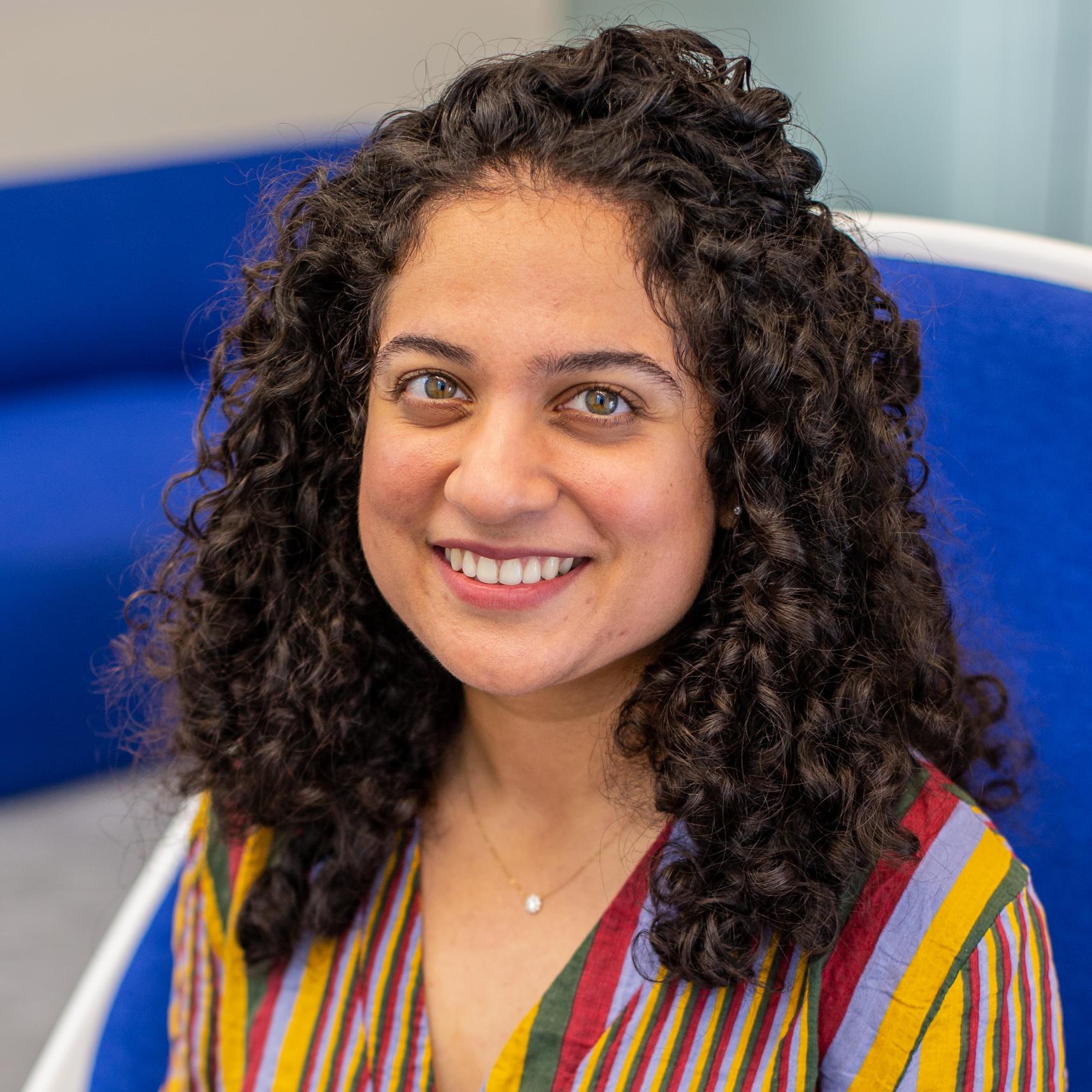 woman with curly dark hair and a brightly patterned shirt sits in a bright blue chair