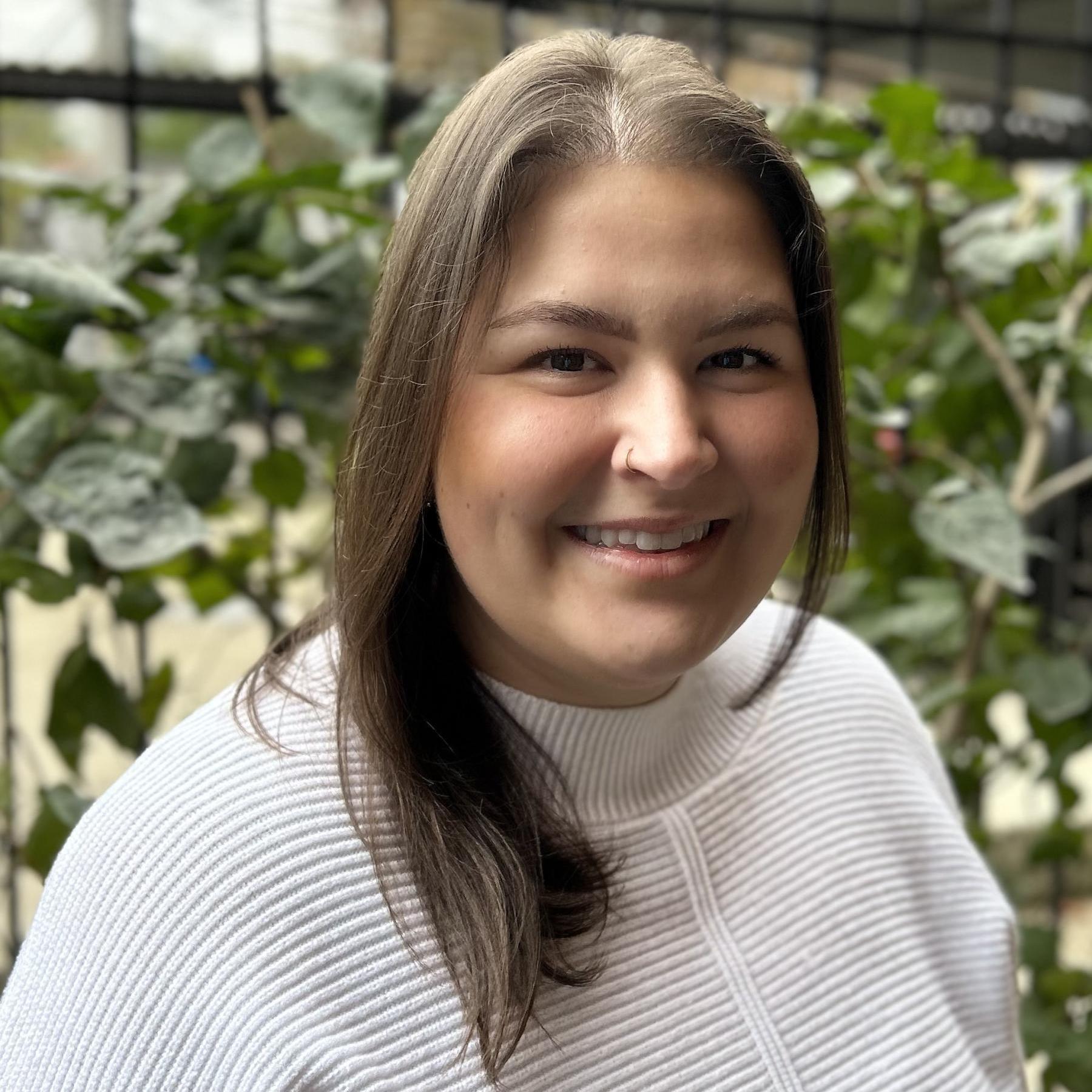 A woman in a white sweater and chest-length brown hair stands in front of plants