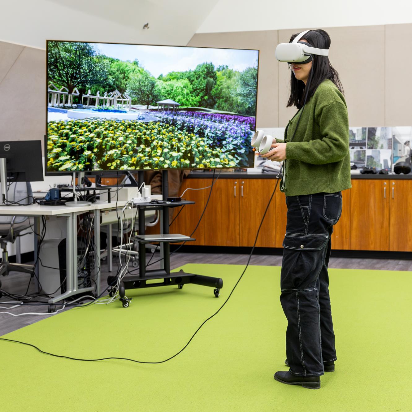 person in a VR headset standing in a room with bright green carpet and a large monitor showing a garden scene