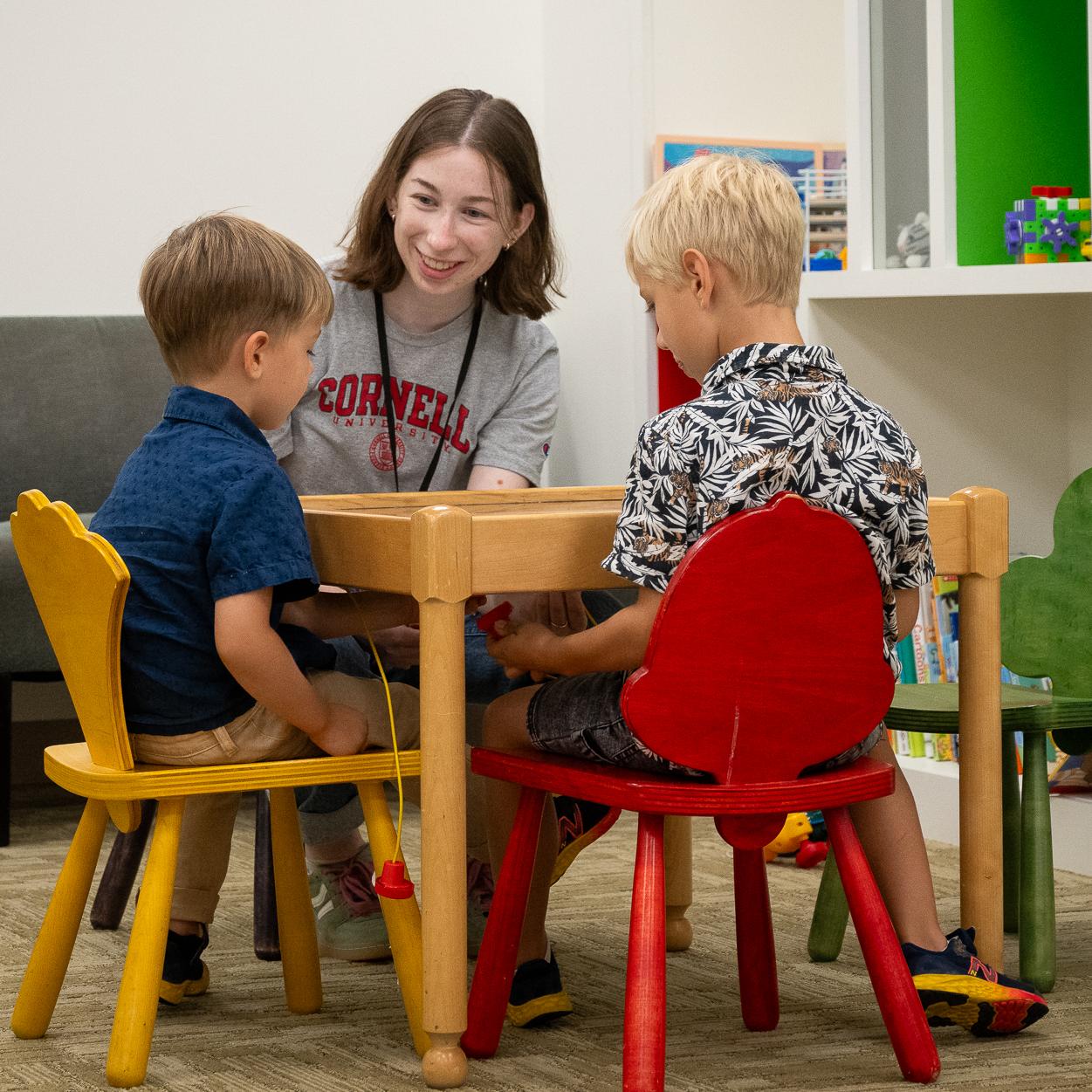 graduate student in a Cornell sweatshirt sits at a colorful child-sized table and chairs talking to two small children in a colorful playroom