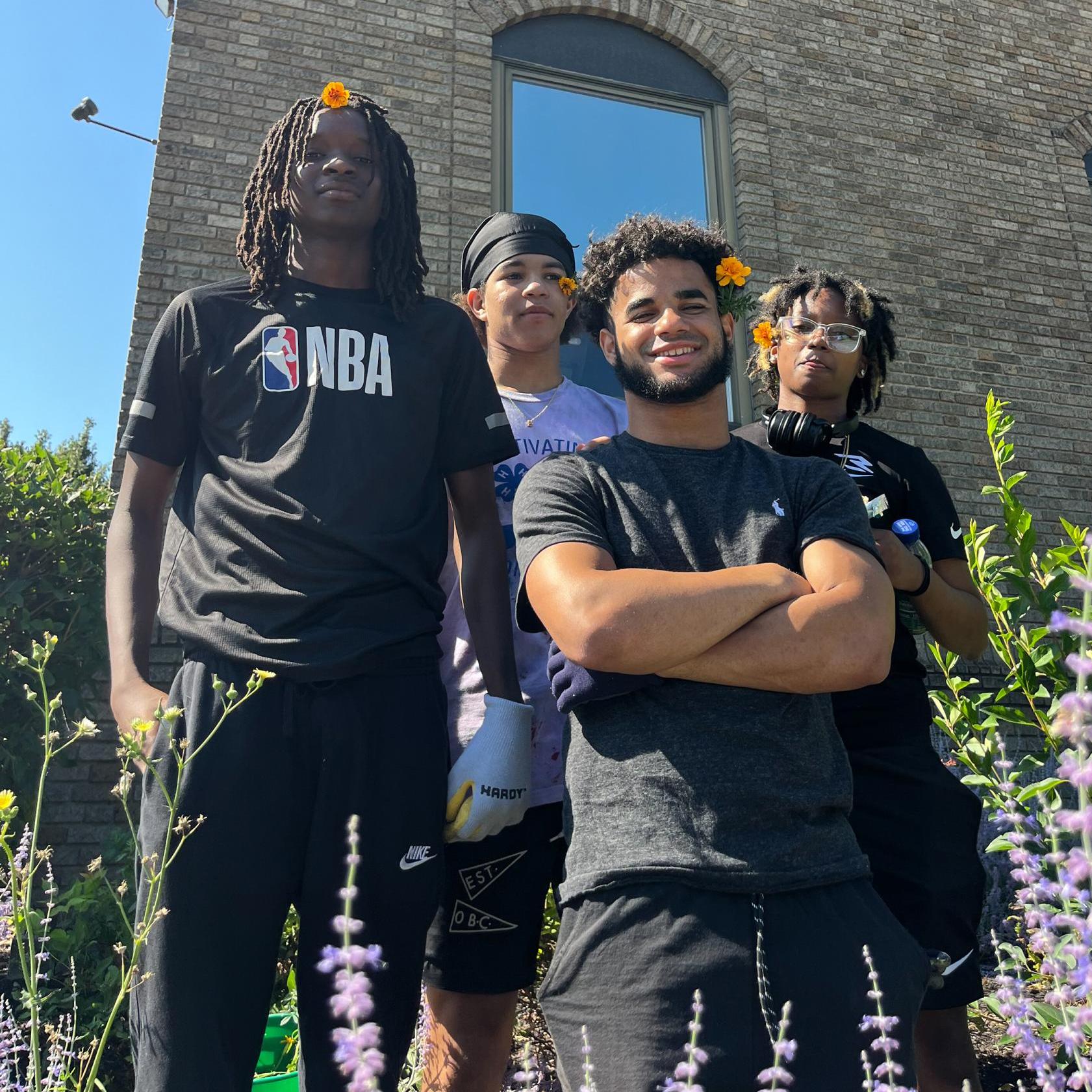 group of four young people standing in a garden with a building behind them. They wear flowers in their hair.