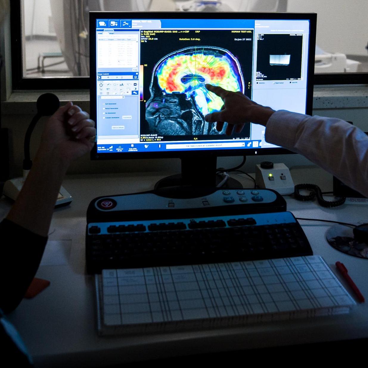 two people in a darkened room look at a brain scan on a monitor. an MRI machine is visible through a window