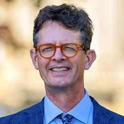 A man with orange glasses, wearing a blue dress shirt, blue paisley tie, and a blue checkered sportcoat.