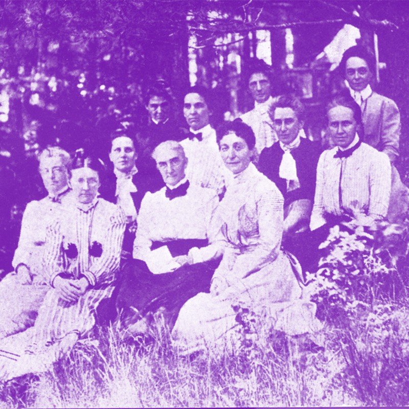 1906 group of faculty sitting on the ground in a wooded area. black and white photo with a purple color wash