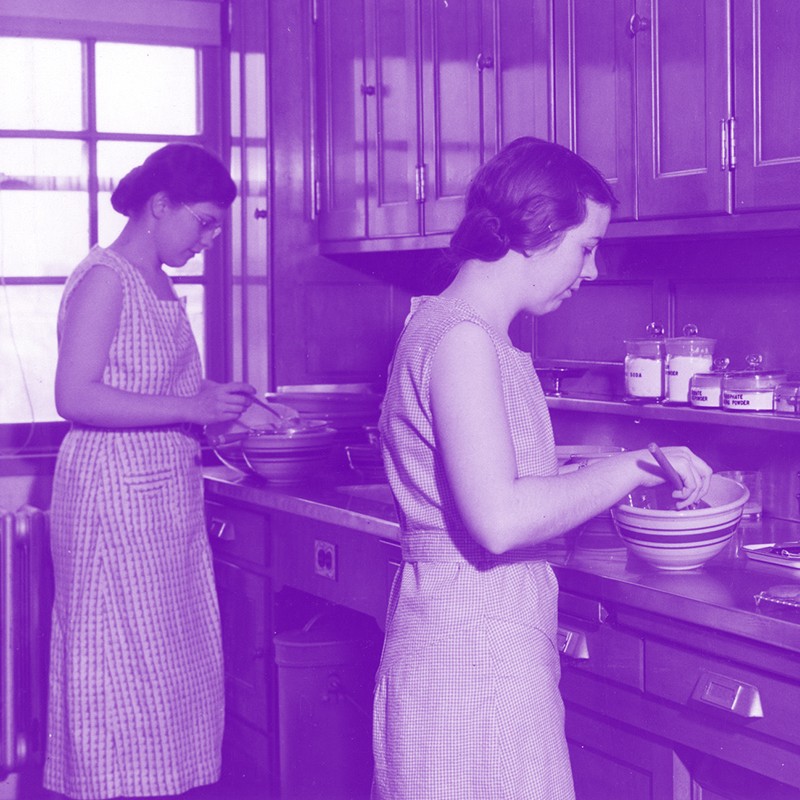 1935 black and white photo tinted purple of two women working in a kitchen