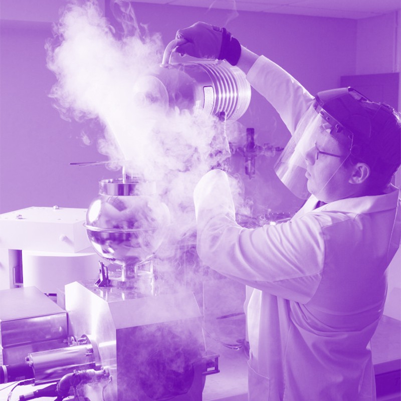 Black and white photo with a purple tint showing a man in a lab coat and face shield pouring liquid nitrogen in a lab setting