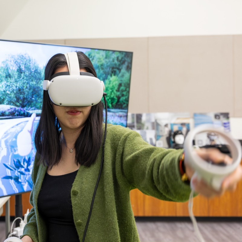 student wearing a virtual reality headset holding out a sensor ahead of her with a large monitor behind her showng a path through some trees