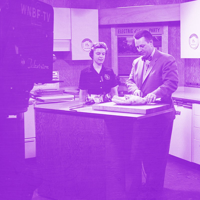 Historical black and white photo with a purple tint showing a woman and man on the set of a TV show doing a cooking demo