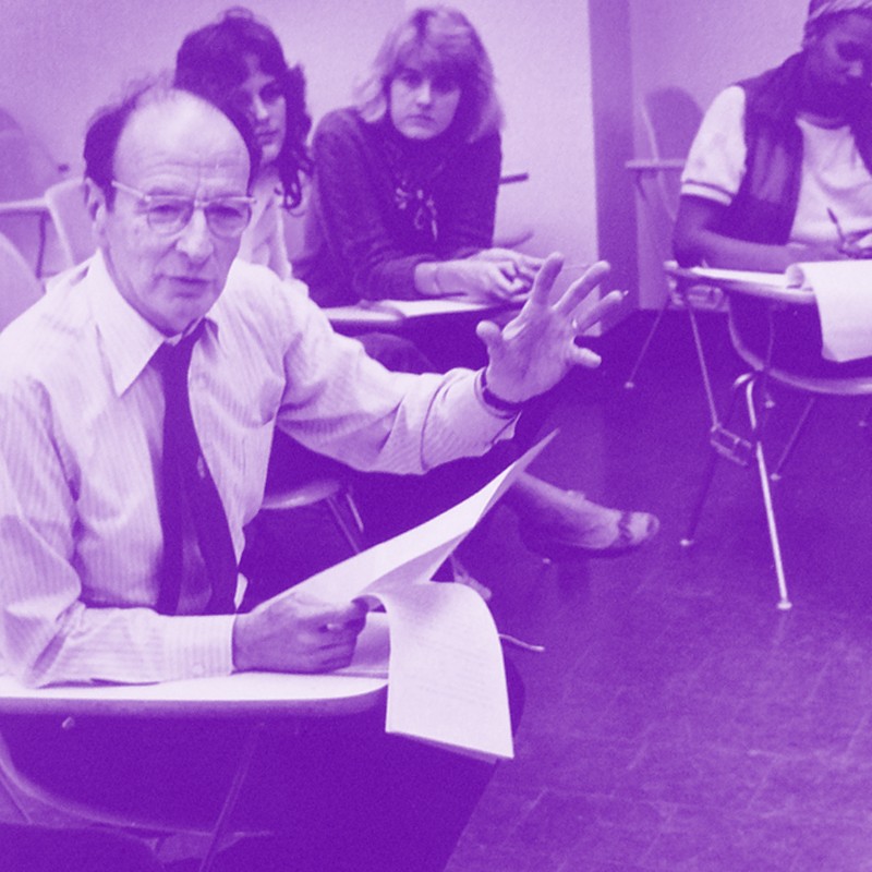 Historical black and white photo with a purple tint of a man speaking in a college classroom